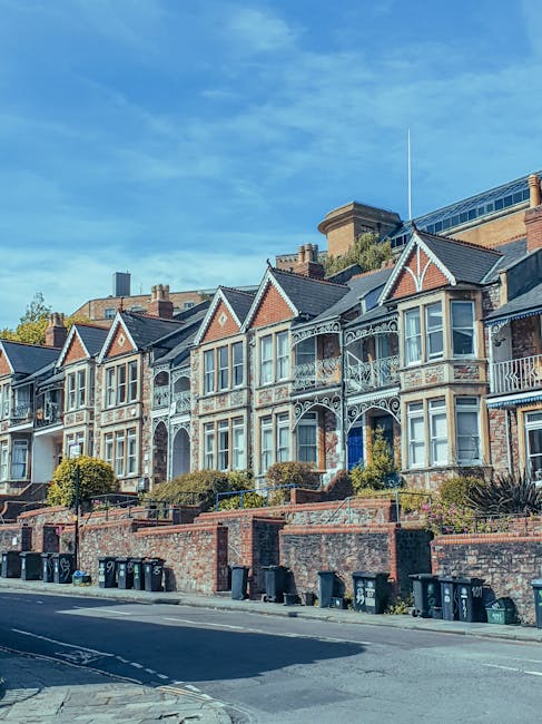 A row of Victorian-style terraced houses with decorative wrought iron balconies and bay windows, situated on a residential street under a clear blue sky. The houses are built with red brick and feature gabled roofs, with some having small front gardens separated by low brick walls. In front of the properties, a paved pavement runs along the street lined with black wheelie bins and some greenery, including neatly trimmed bushes. The scene captures a typical urban residential environment suitable for a house removal or local home relocation service, with no visible moving equipment or personnel present in the image. The image is relevant to the services offered by Removal Van Bermondsey, illustrating the type of houses often involved in local removals and furniture transport within Bermondsey.