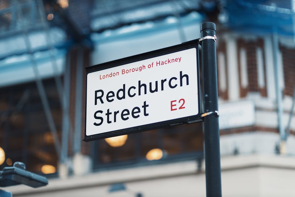 A street sign displaying 'Redchurch Street' in bold black letters with a red 'E2' postcode on a black and white background. The sign is mounted on a black pole situated on the pavement, with part of a modern building with large glass windows and a blue metal structure visible behind it. The image shows the sign during daytime with soft lighting and some blurred architectural details in the background. This scene illustrates a typical urban environment in Bermondsey, London, relevant to house removals and home relocation services offered by Removal Van Bermondsey, capturing the context of moving logistics and street-based furniture transport.